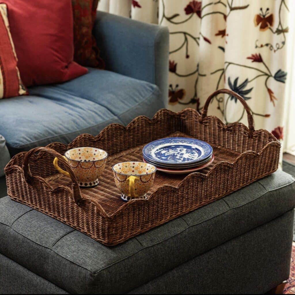 Scalloped teak rattan serving tray with handles styled on a grey upholstered ottoman with patterned cups and plates in a cosy living room.