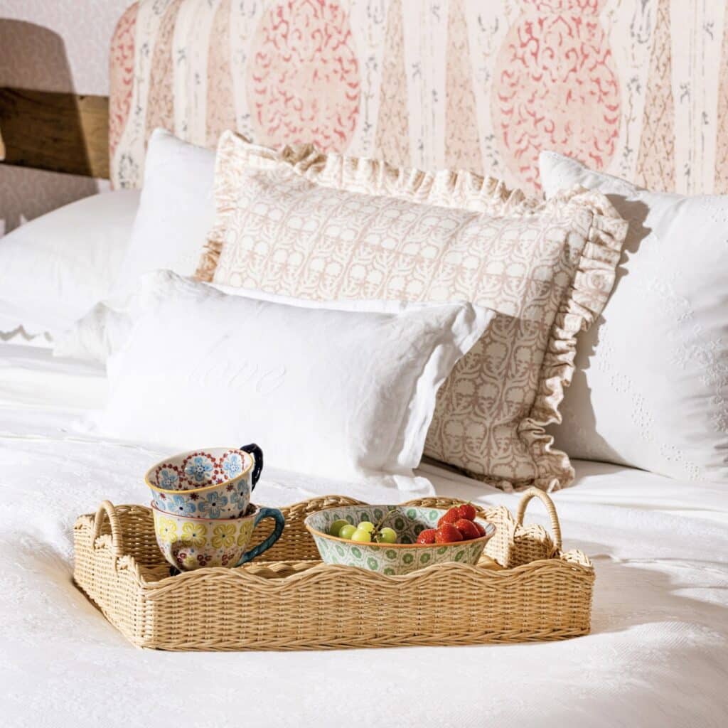 Scalloped rattan breakfast tray on a bed with patterned cups, fruit and cushions behind.