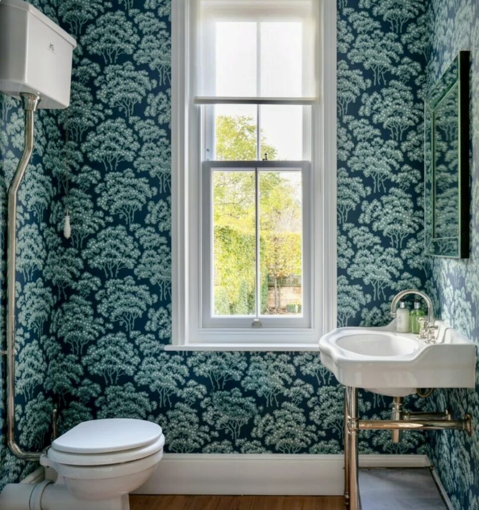 Green floral wallpaper in bathroom with vintage sink and sash window.