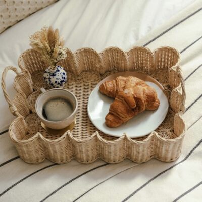 Scalloped wicker breakfast tray with artisan coffee, croissants and dried flowers on a striped bedspread