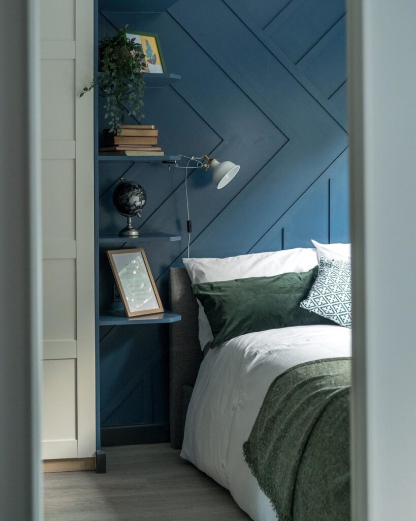 Floating bedside shelves in Farrow & Ball Stiffkey Blue panelled bedroom — wall-mounted shelving beside bed with lamp, books and globe replacing a bedside table