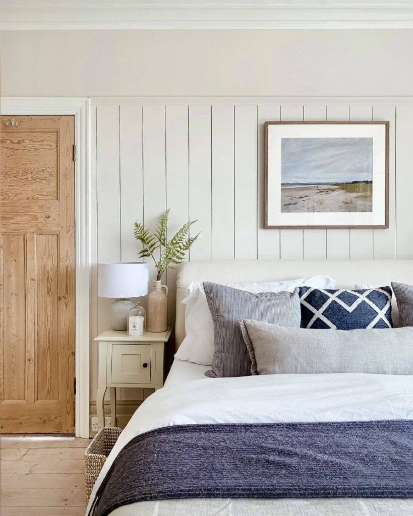 Relaxed coastal interior bedroom painted in Farrow & Ball School House White and Drop Cloth with layered linen bedding, navy cushions and a natural wood door