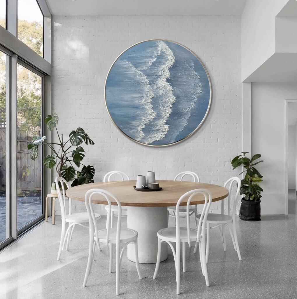 Round wave painting in a modern dining area with white chairs and indoor plants, styled with coastal interior design elements.