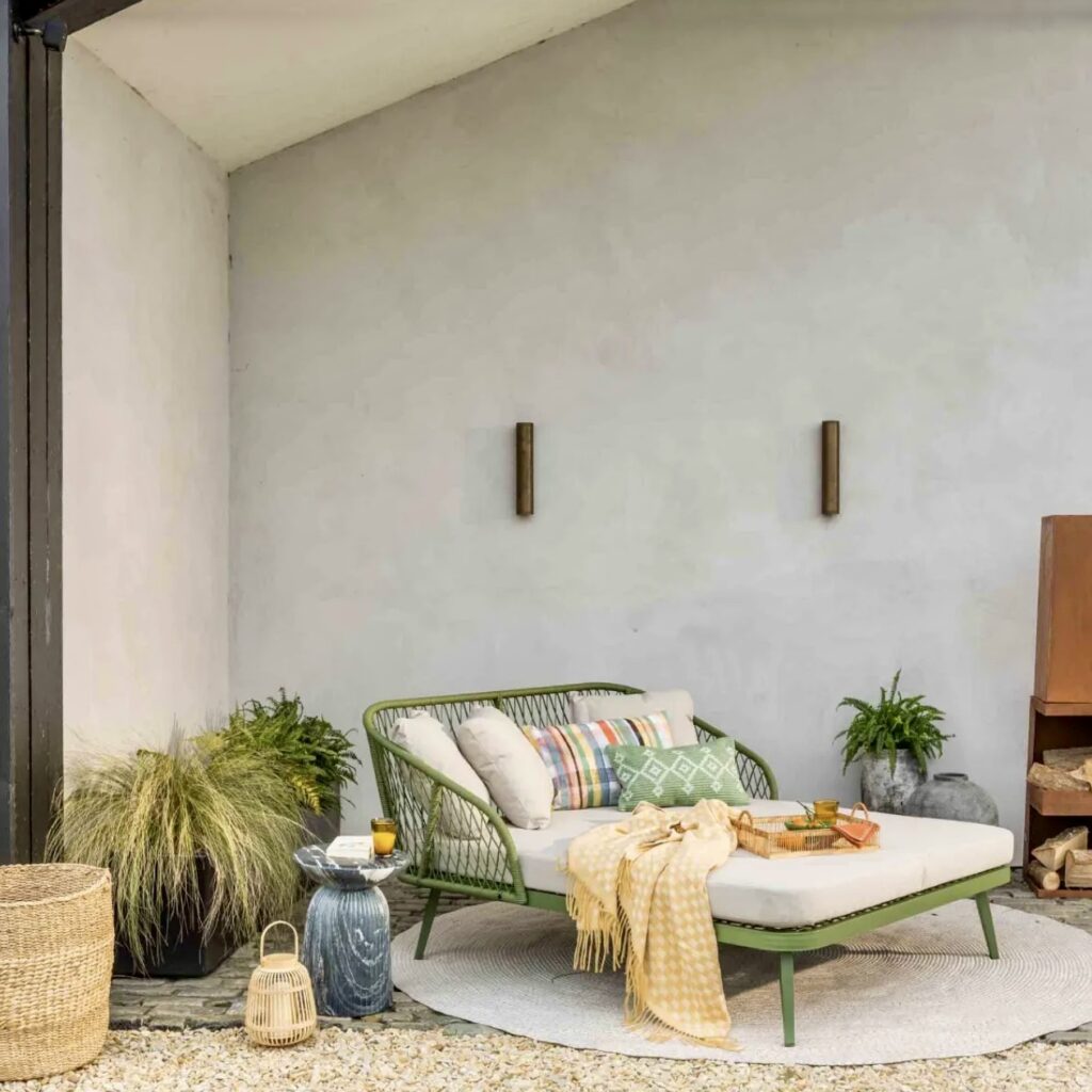 Green woven daybed from Atkin and Thyme styled on gravel with grasses, cushions and lanterns – gravel garden ideas with pots