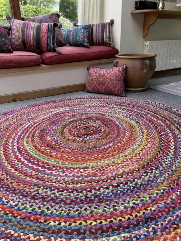 Circular rag rug in bright rainbow colours under a window seat with boho cushions, handmade from recycled fabric.