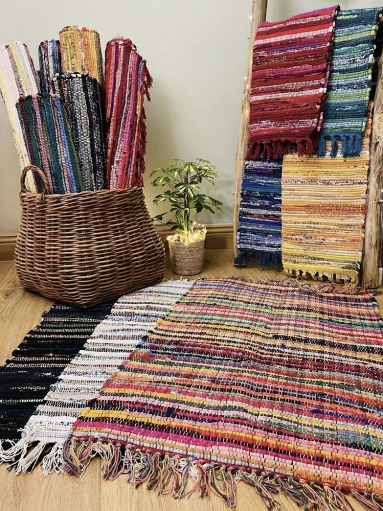 Handwoven rag rug with multicoloured stripes on wood floor, surrounded by rolled recycled rugs and upcycled fabric mats in vibrant hues.