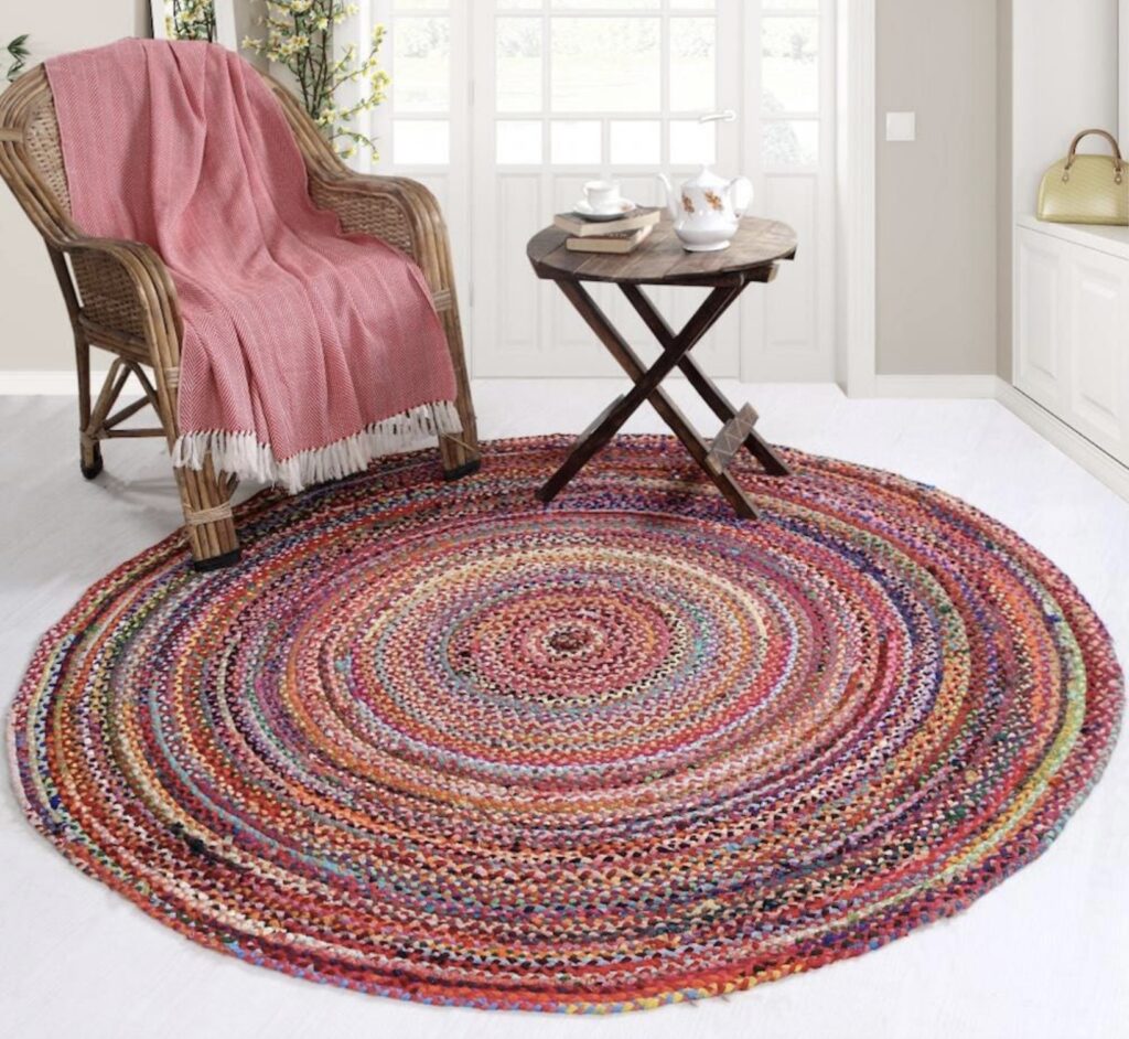 Round braided rag rug in vibrant colours styled with a wicker chair and tea table in a bright sunroom.