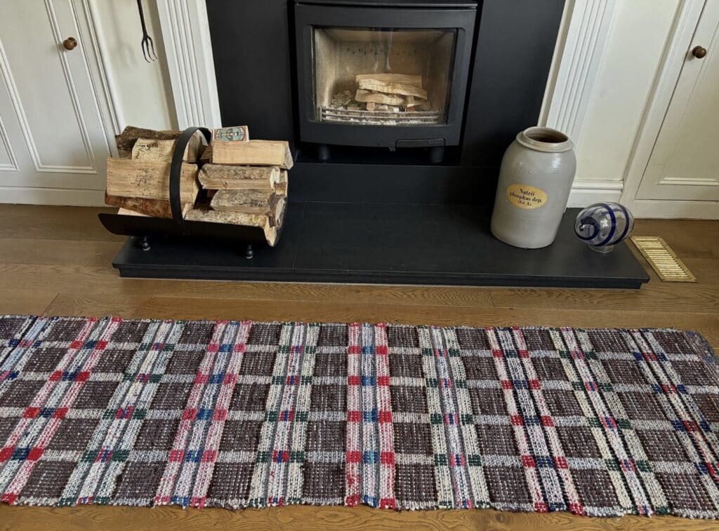 Vintage Swedish handwoven rag rug with red and blue check pattern on wooden floor in front of a black fireplace