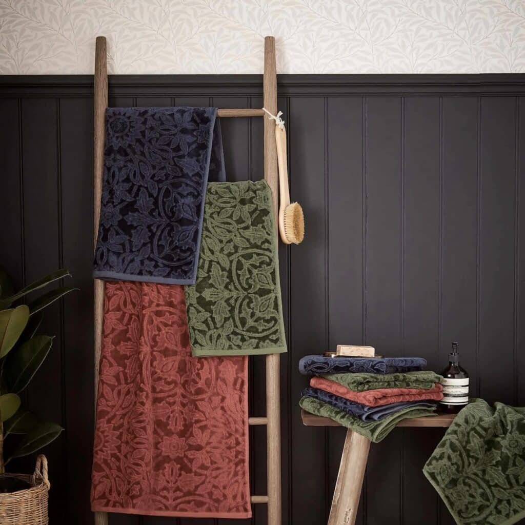 Morris & Co St James towels in red and green draped over a wooden ladder in a dark panelled bathroom