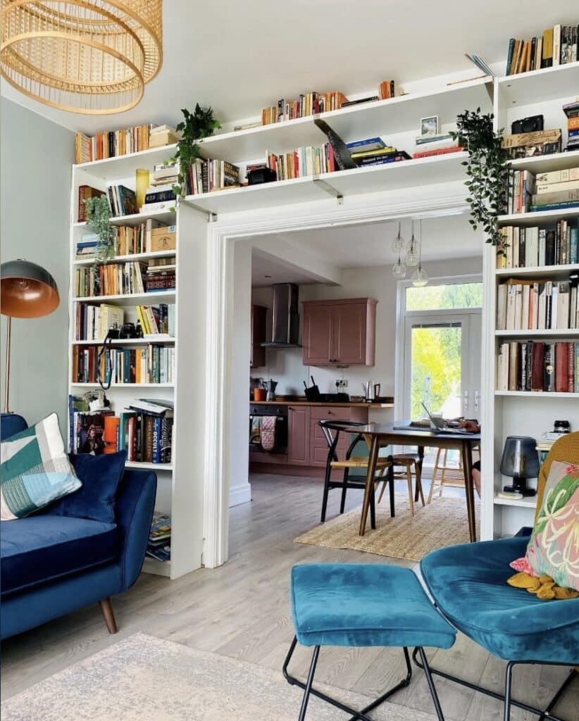 Built-in bookshelves around a doorway in a small living room with storage up high, opening through to a pink kitchen and dining area beyond