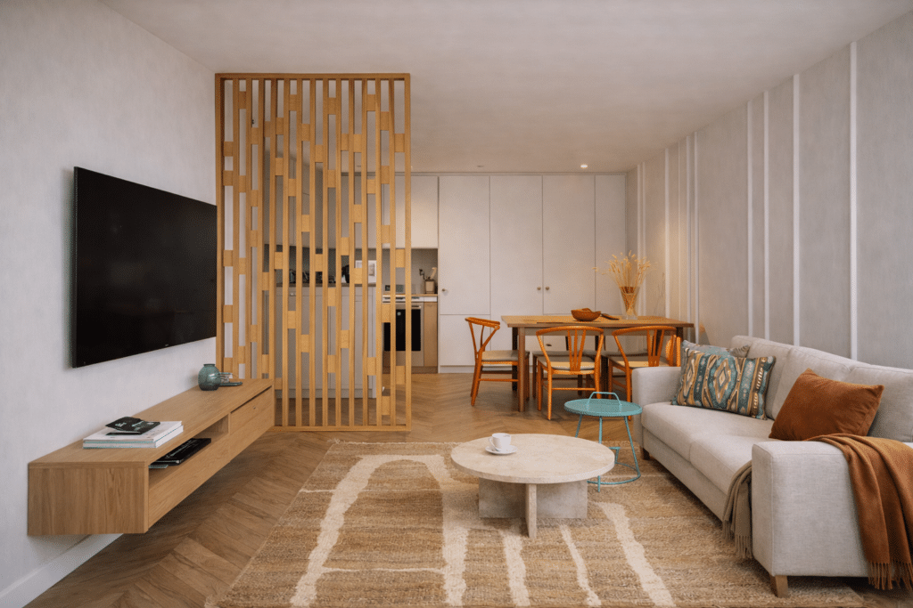 AI visualisation of an open-plan small living room with beige sofa, beige living room rug, slatted oak room divider, wall-mounted TV, orange dining chairs and aqua side table.
