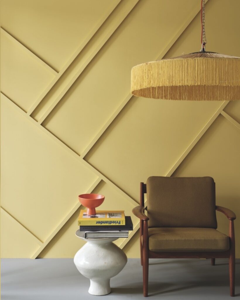 Graphic panelling painted in Farrow & Ball Citrona (mustard yellow) with a fringed pendant light, mid-century armchair and sculptural side table for modern retro living room colour ideas.