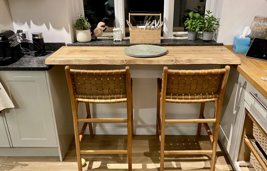 Live edge wooden breakfast bar with two woven bar stools, herbs on the windowsill and a coffee machine in a small UK kitchen.