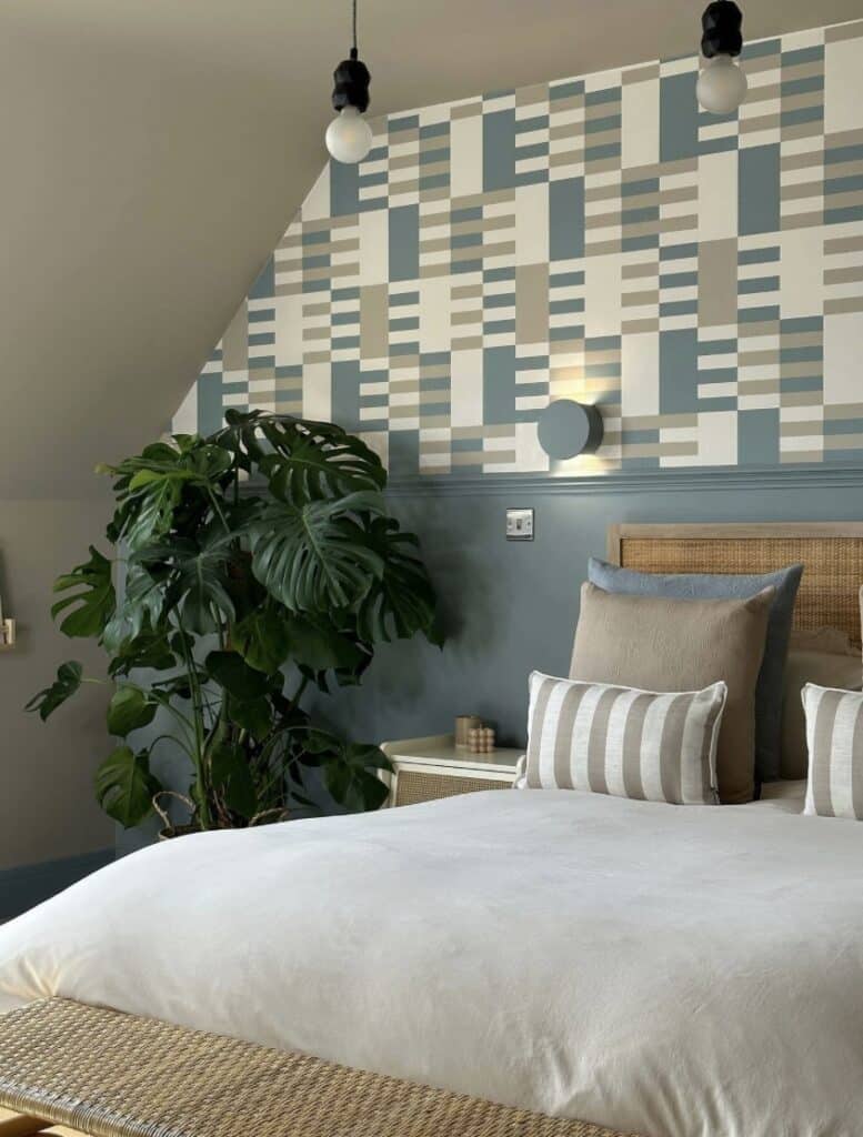 Geometric bedroom wallpaper on a sloped wall above blue-grey panelling, with neutral bedding, rattan bench and a large leafy plant