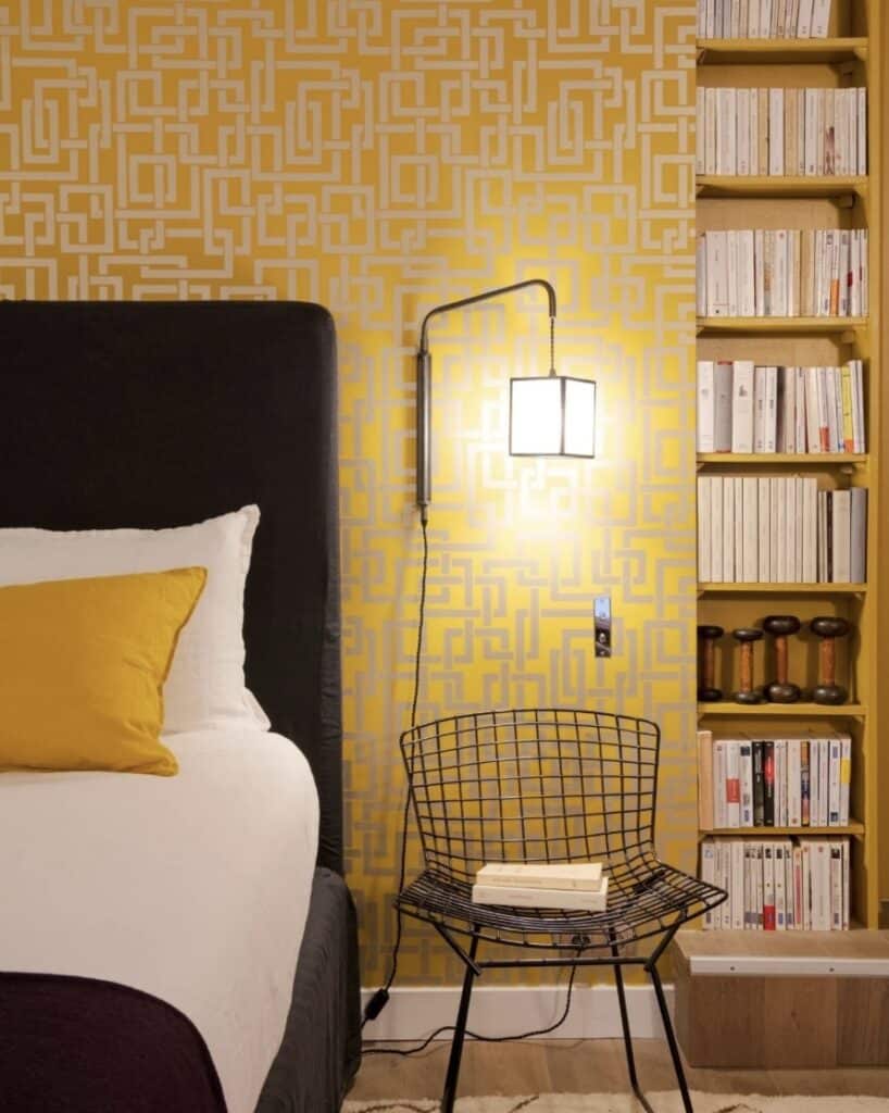 Mustard geometric wallpaper feature wall behind a bed with a dark upholstered headboard, white bedding and a black chair