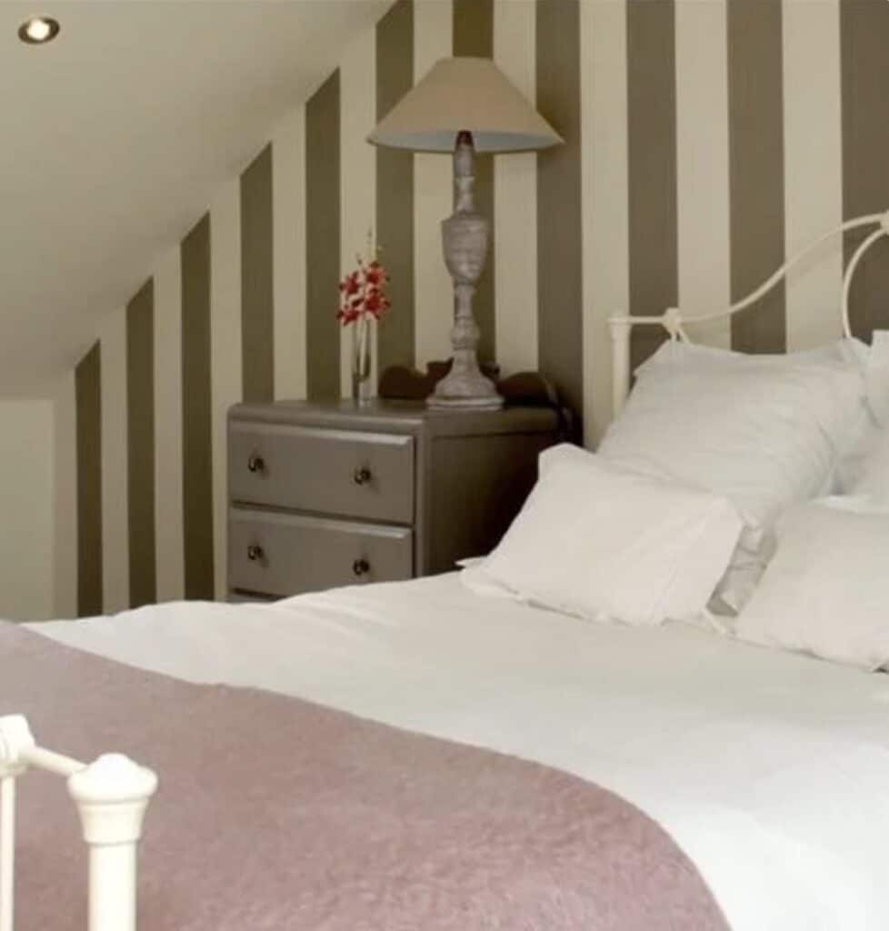 Broad Stripe Wallpaper by Farrow & Ball in warm beige and cream stripes behind a white bed – calming bedroom wallpaper ideas for beige bedroom ideas.