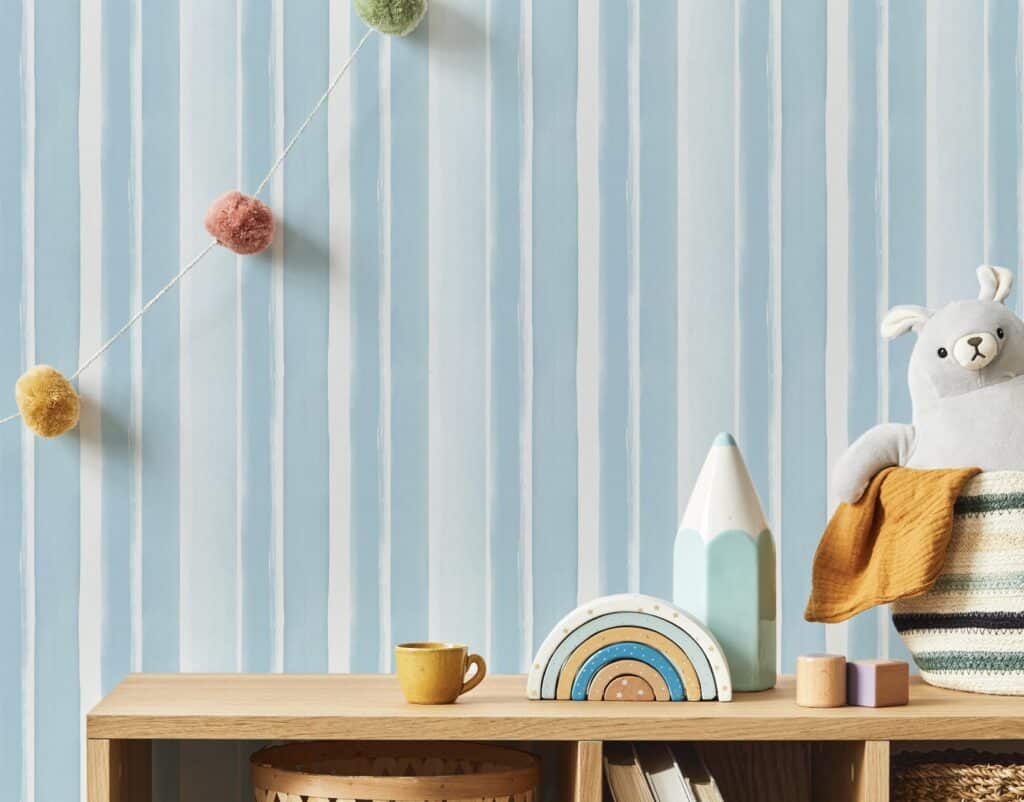 Pale blue striped wallpaper behind a wooden shelf with rainbow toy, teddy and pom-pom garland – calm nursery décor and blue bedroom ideas.