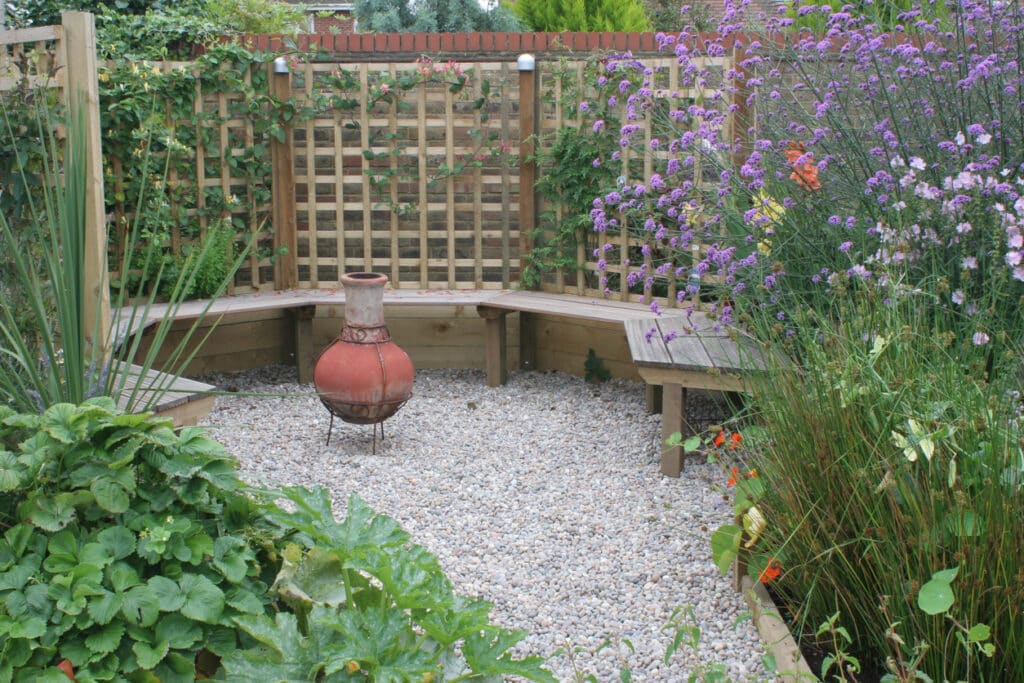 Small garden ideas: curved built-in timber bench with gravel seating area and chimney pot, surrounded by cottage-style planting