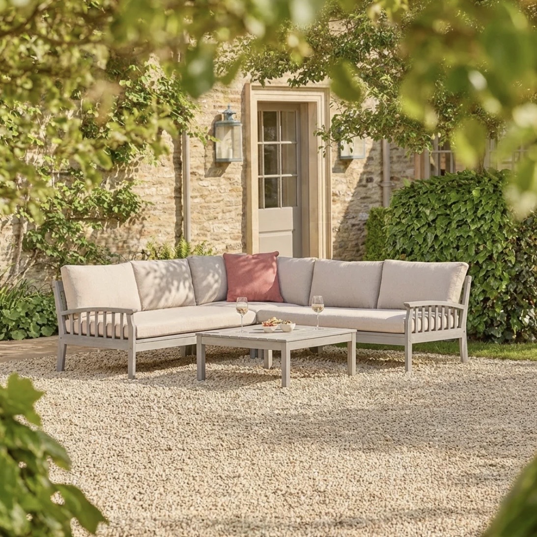 Wooden corner garden sofa with pale cushions on gravel in a leafy garden