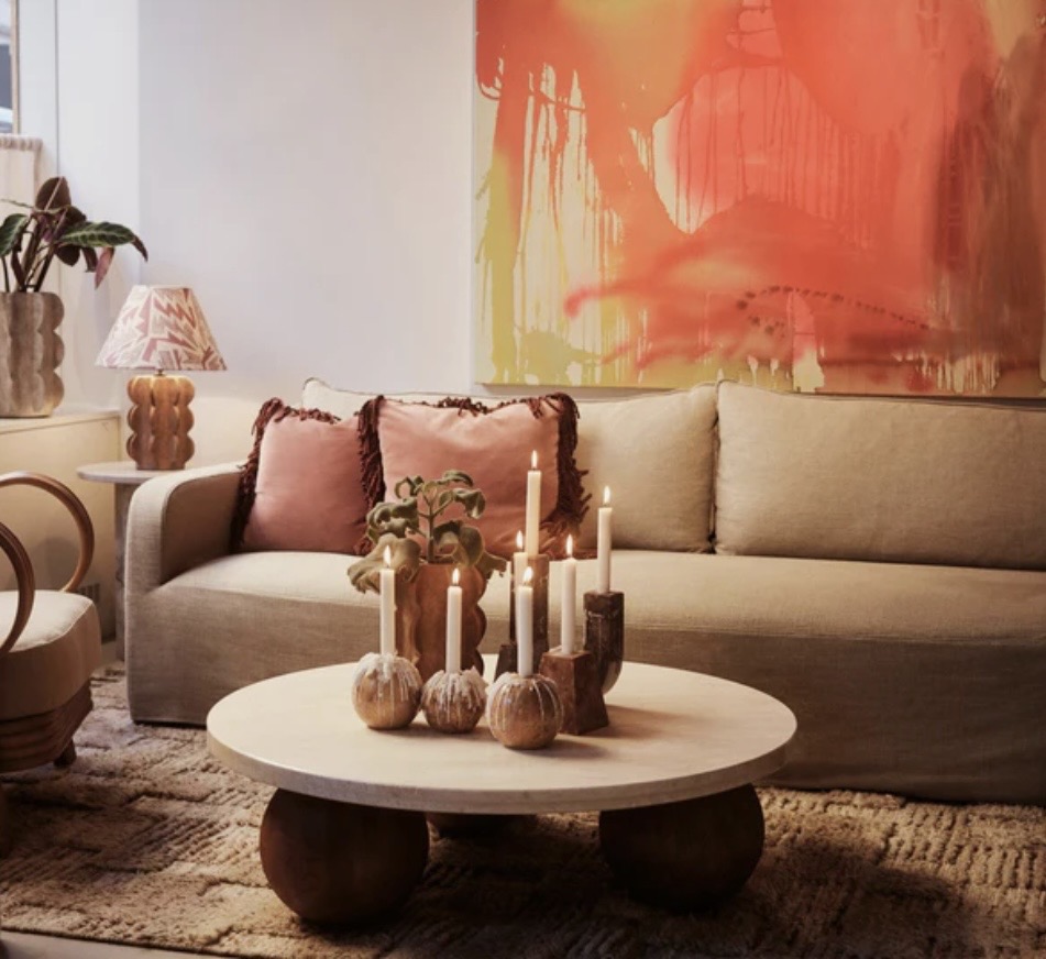 Grouped candles styled on a round coffee table in a warm living room with blush cushions and bold artwork, showing soft, atmospheric coffee table styling.