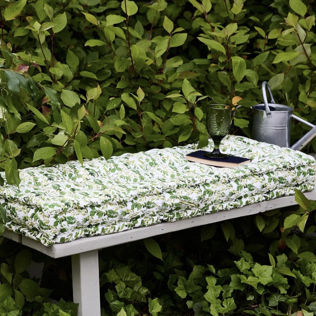 Botanical leaf print outdoor bench cushion in white and green, 100% cotton, styled on grey garden bench with lush green foliage backdrop