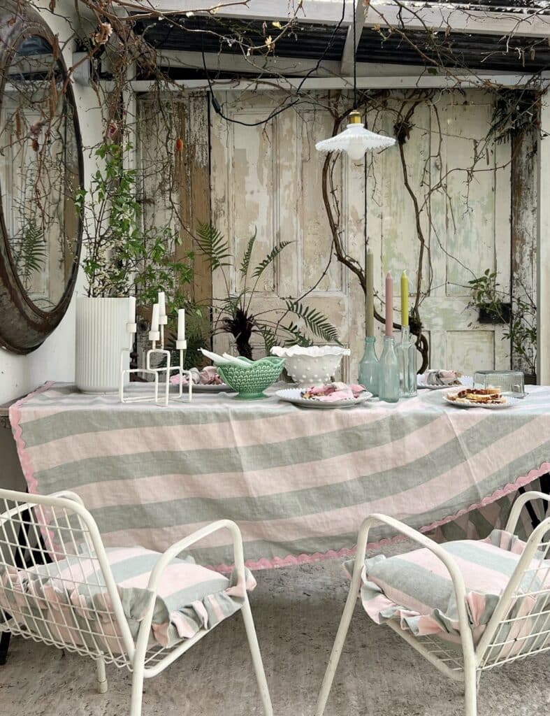 Striped linen outdoor cushion cover in soft pastel tones of blush, sage and cream, styled on garden seating with trailing foliage and candlesticks