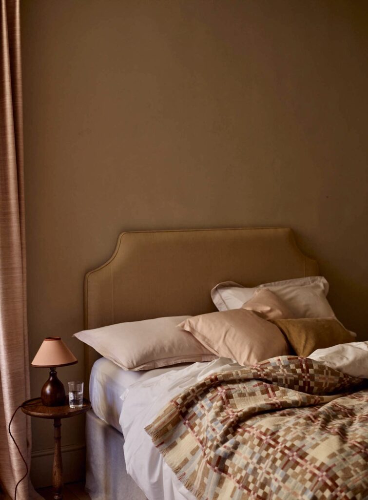 Pink and beige bedroom with a straw cotton upholstered headboard, blush pillowcases, a pink lampshade and a woven check blanket against brown-beige walls