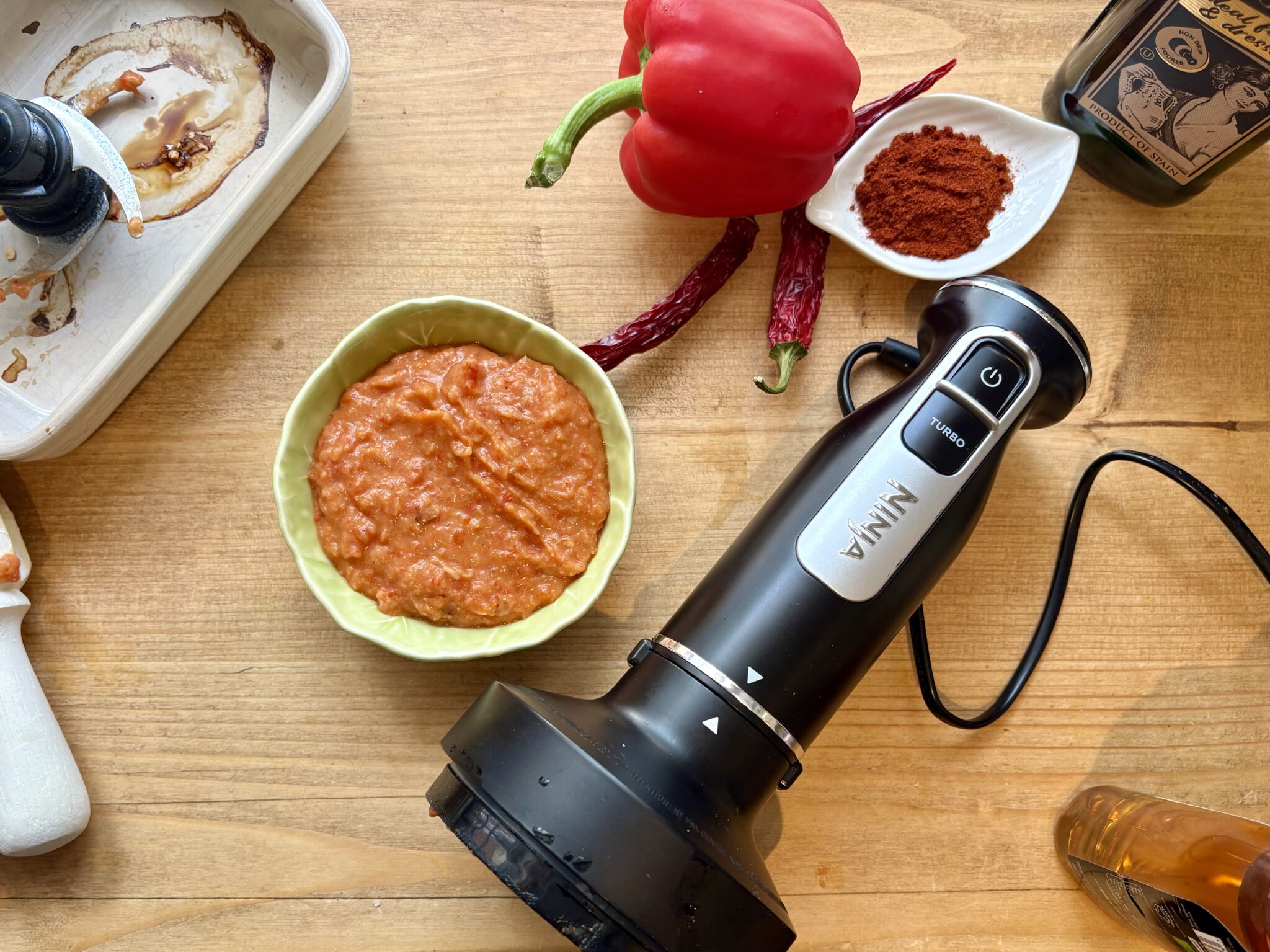 Ninja Foodi hand blender next to roasted red pepper dip in a green bowl, with smoked paprika, red pepper and dried chillies on a wooden chopping board