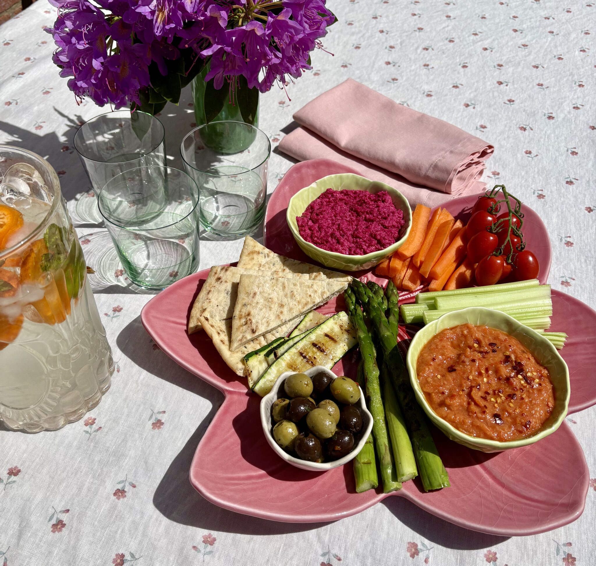 Roasted red pepper dip in a green bowl on a pink flower plate with beetroot hummus, grilled asparagus and courgette, flatbreads, olives, carrots and cherry tomatoes, styled outdoors with rhododendrons and green glasses