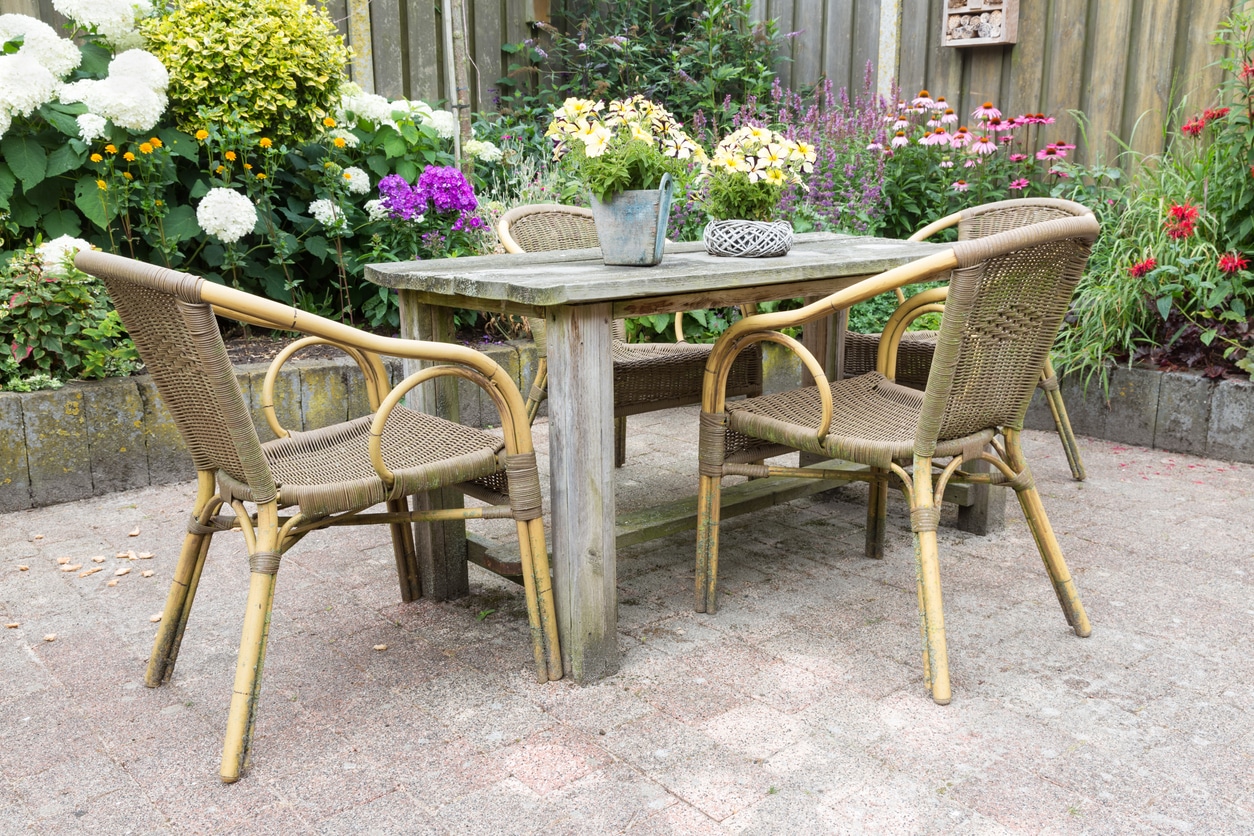 Cottage small garden ideas: weathered wooden dining table with rattan chairs, packed cottage planting border and insect hotel on timber fence