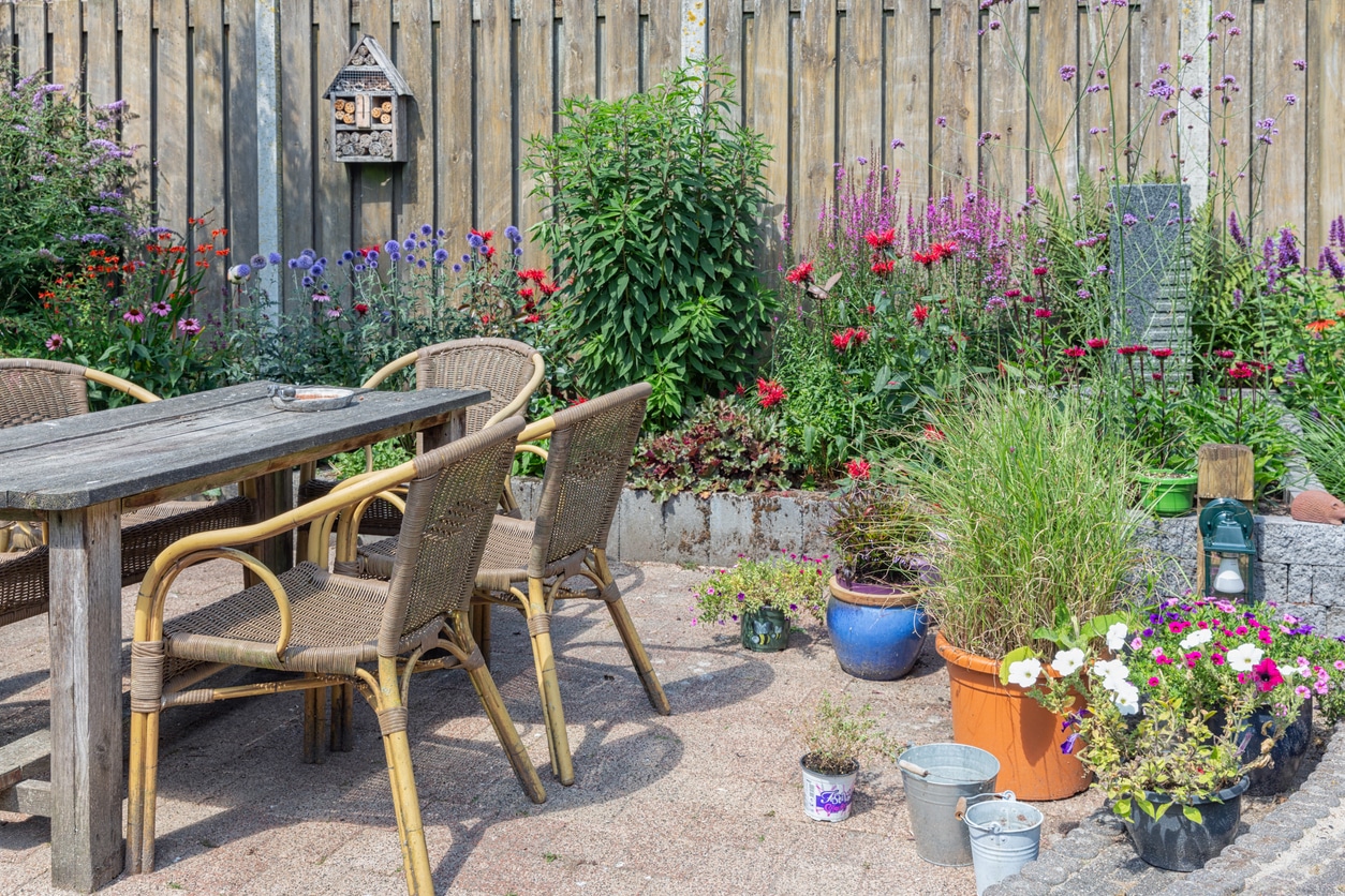 Cottage small garden ideas: rattan bistro chairs and wooden table on granite paving with deep cottage planting border, pots and lanterns