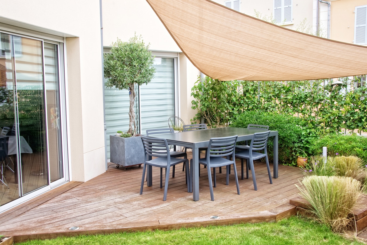 Small garden decking ideas: modern timber deck with dining table and chairs, sail shade overhead and olive tree in planter