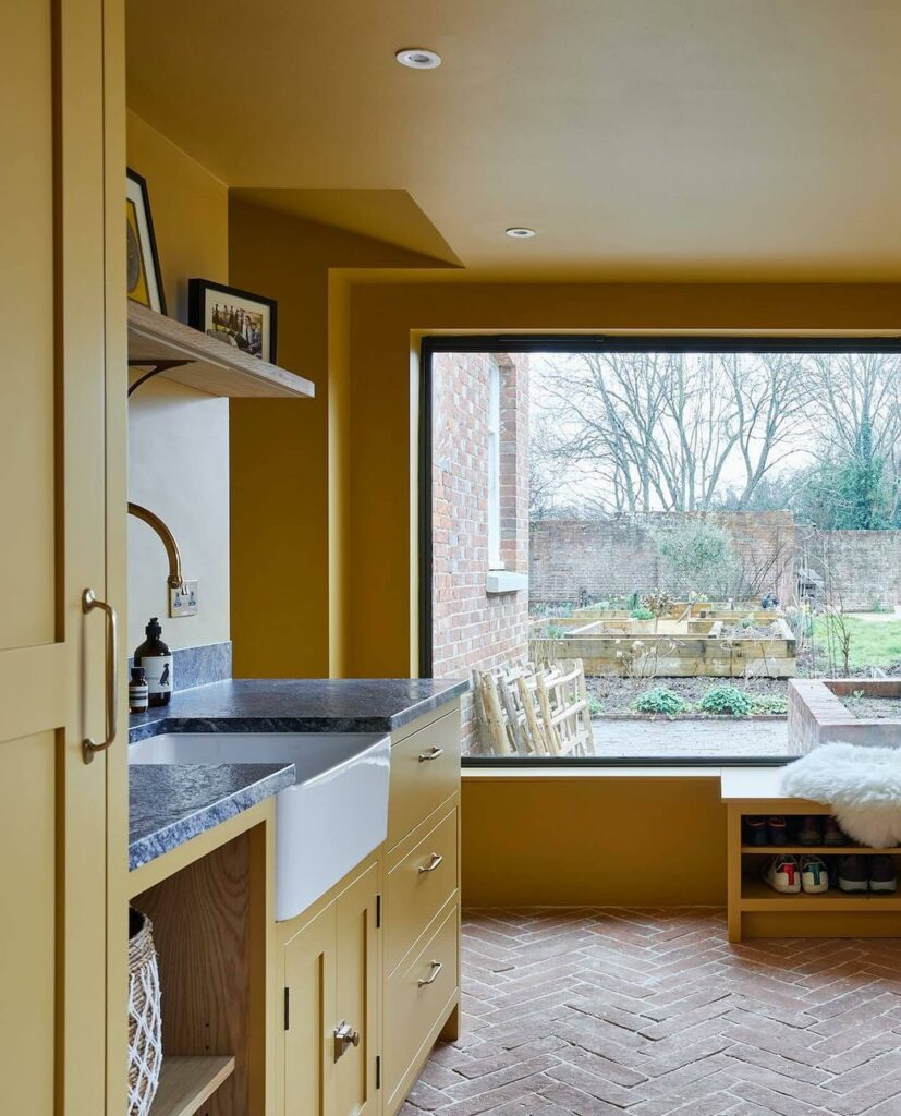 Warm yellow kitchen painted in Farrow & Ball India Yellow with farmhouse sink, dark stone worktops and herringbone brick flooring