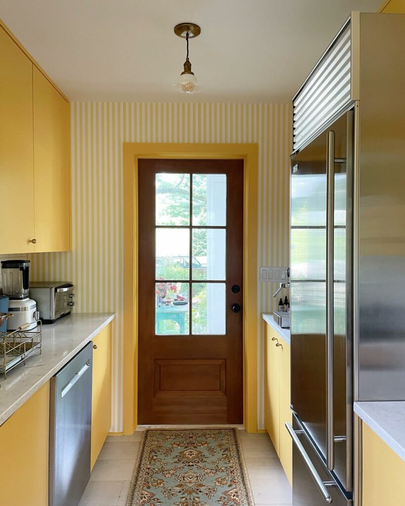 Bright yellow galley kitchen painted in Farrow & Ball Babouche with striped wallpaper, stainless steel appliances and a vintage runner rug