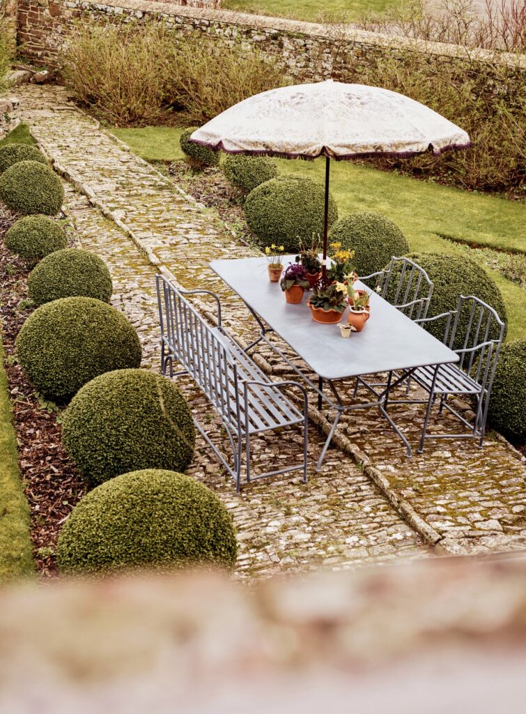 Rowen & Wren Ludlow Rectangular garden dining table on a cobbled terrace with topiary balls and vintage parasol — outdoor dining ideas
