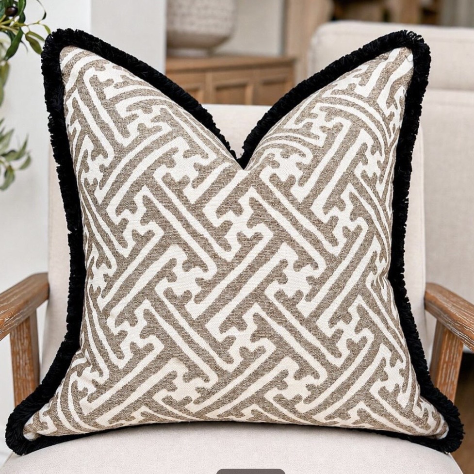 Beige and taupe geometric cushion with black fringe trim on a neutral armchair