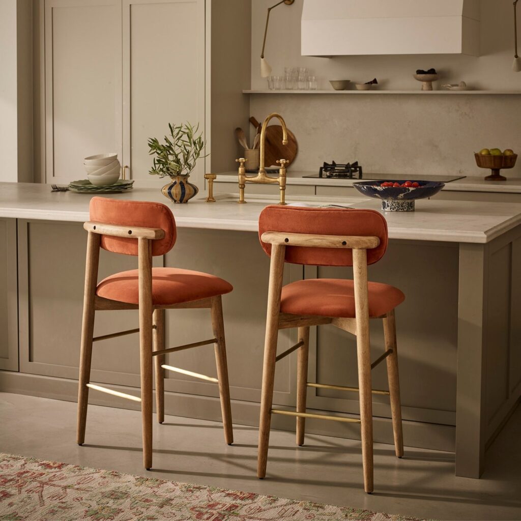 Two rust velvet bar stools with oak legs and brass footrails at a stone kitchen island