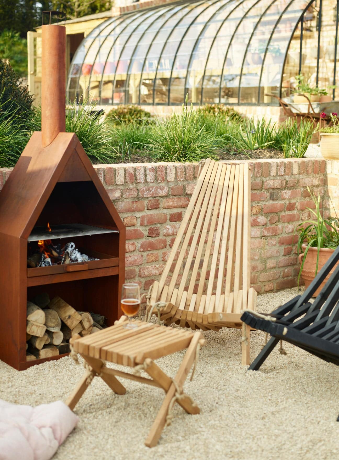 Harmen birch outdoor chair beside a lit corten fire oven and stacked logs on a gravel terrace
