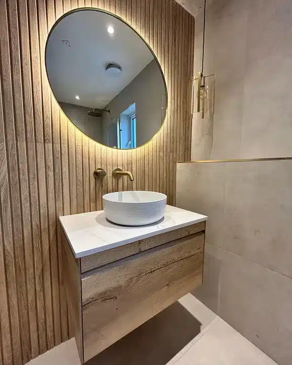 Vertical wood-effect tile feature wall behind a vessel basin with a backlit mirror and brass taps