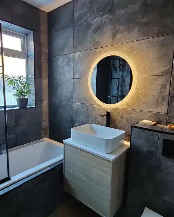 Family bathroom with large dark slate-effect wall tiles, light oak vanity and backlit round mirror