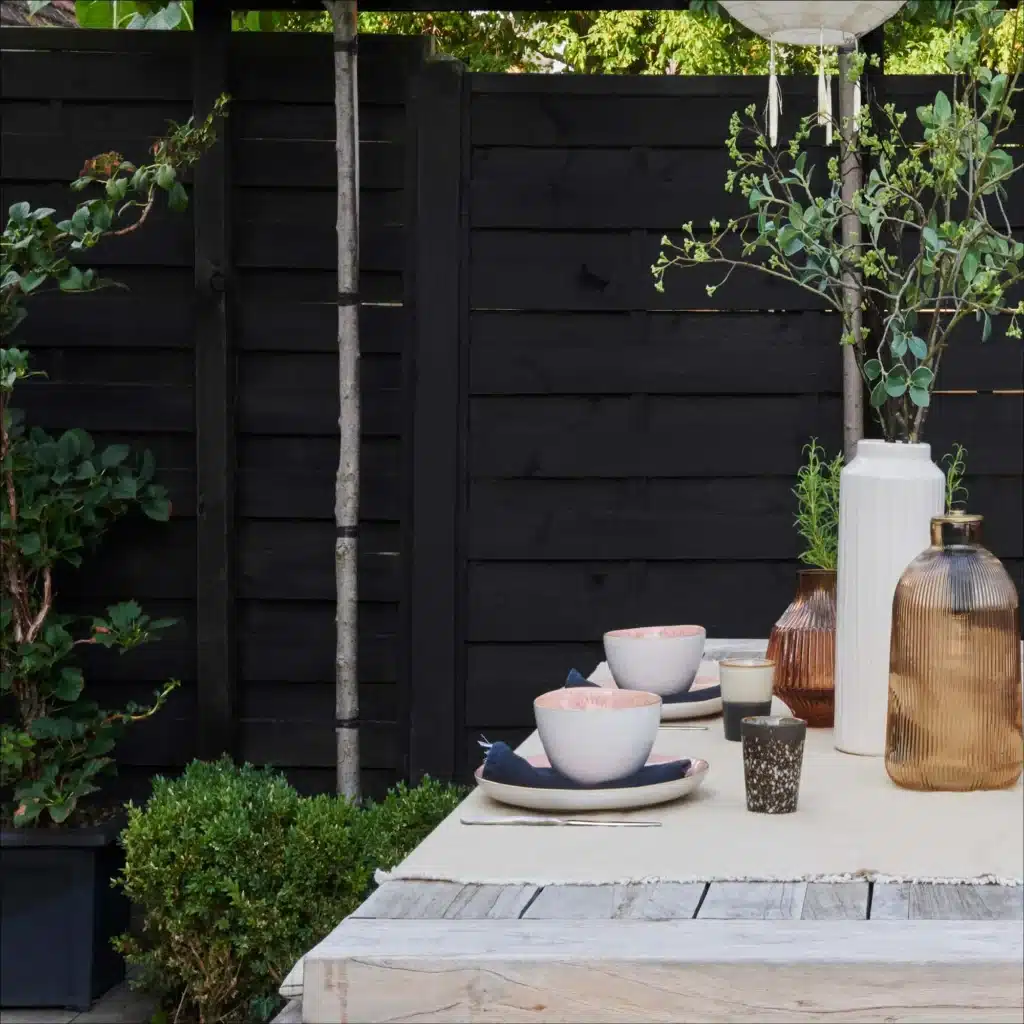 Horizontal fence boards painted matt black behind an outdoor dining table set with pink bowls and amber glass vases