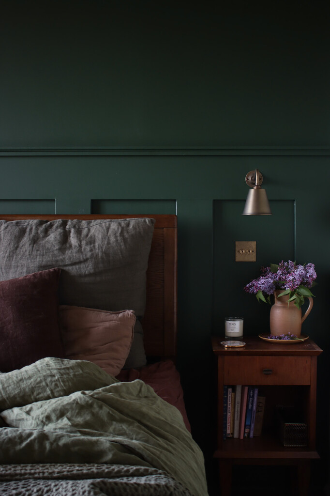 Forest green bedroom with Duck Green panelled walls, a brass wall light, a wooden bed with linen bedding and lilac in a terracotta jug