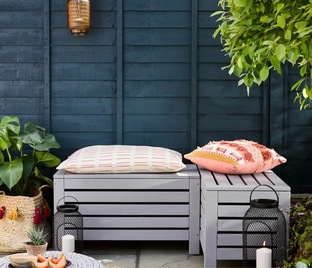 Overlapping fence panels painted deep navy blue behind grey storage benches with coral cushions and black lanterns
