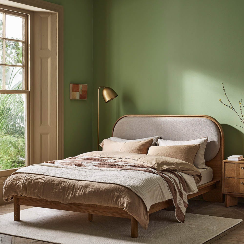 Green and cream bedroom with olive-green walls, a wooden bed with an upholstered headboard, layered neutral bedding and a brass wall light.